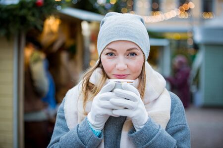 Young woman in Christmas market drinking cup of hot chocolate with marshmallow wearing knitted warm hat and scarf. Illuminated and decorated fair kiosks and shops on background. Helsinki, Finlandの写真素材