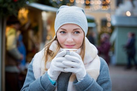 Young woman in Christmas market drinking cup of hot chocolate with marshmallow wearing knitted warm hat and scarf. Illuminated and decorated fair kiosks and shops on background. Helsinki, Finlandの写真素材