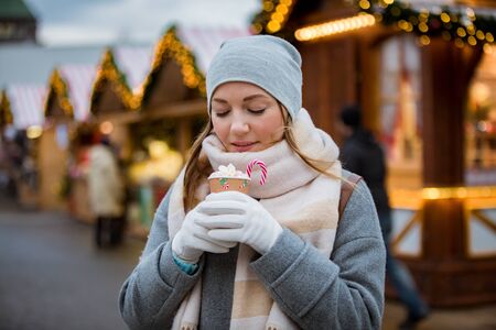 Young woman in Christmas market drinking cup of hot chocolate with marshmallow wearing knitted warm hat and scarf. Illuminated and decorated fair kiosks and shops on background. Helsinki, Finlandの写真素材