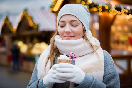 Young woman in Christmas market drinking cup of hot chocolate with marshmallow wearing knitted warm hat and scarf. Illuminated and decorated fair kiosks and shops on background. Helsinki, Finlandの写真素材