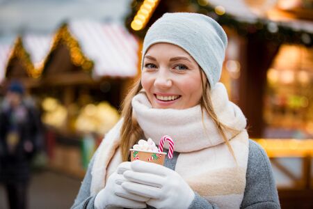 Young woman in Christmas market drinking cup of hot chocolate with marshmallow wearing knitted warm hat and scarf. Illuminated and decorated fair kiosks and shops on background. Helsinki, Finlandの写真素材