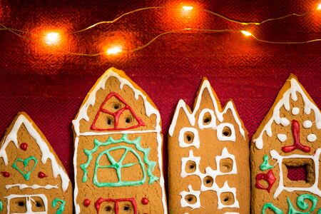 Christmas background. Red table cloth with town of cute gingerbread houses decorated with icing, Christmas lights, glitter and fir tree branches. Holiday mood.の写真素材