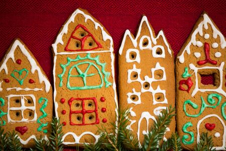 Christmas background. Red table cloth with town of cute gingerbread houses decorated with icing, Christmas lights, glitter and fir tree branches. Holiday mood.の写真素材