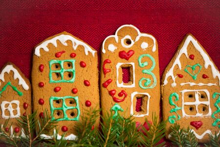 Christmas background. Red table cloth with town of cute gingerbread houses decorated with icing, Christmas lights, glitter and fir tree branches. Holiday mood.の写真素材