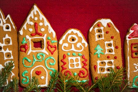 Christmas background. Red table cloth with town of cute gingerbread houses decorated with icing, Christmas lights, glitter and fir tree branches. Holiday mood.の写真素材
