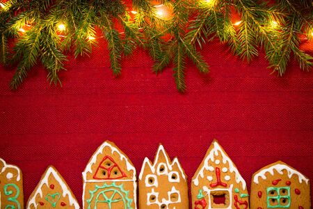 Christmas background. Red table cloth with town of cute gingerbread houses decorated with icing, Christmas lights, glitter and fir tree branches. Holiday mood.の写真素材