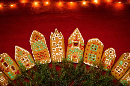 Christmas background. Red table cloth with town of cute gingerbread houses decorated with icing, Christmas lights, glitter and fir tree branches. Holiday mood.の写真素材