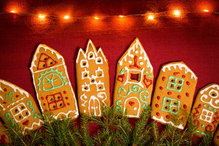 Christmas background. Red table cloth with town of cute gingerbread houses decorated with icing, Christmas lights, glitter and fir tree branches. Holiday mood.の写真素材