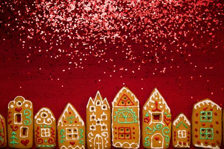 Christmas background. Red table cloth with town of cute gingerbread houses decorated with icing, Christmas lights, glitter. Holiday mood.の写真素材