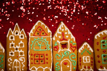 Christmas background. Red table cloth with town of cute gingerbread houses decorated with icing, Christmas lights, glitter. Holiday mood.の写真素材