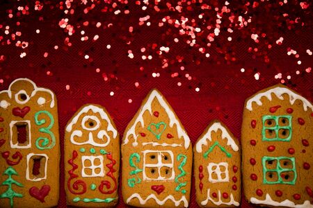 Christmas background. Red table cloth with town of cute gingerbread houses decorated with icing, Christmas lights, glitter. Holiday mood.の写真素材