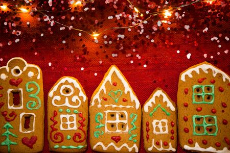 Christmas background. Red table cloth with town of cute gingerbread houses decorated with icing, Christmas lights, glitter. Holiday mood.の写真素材