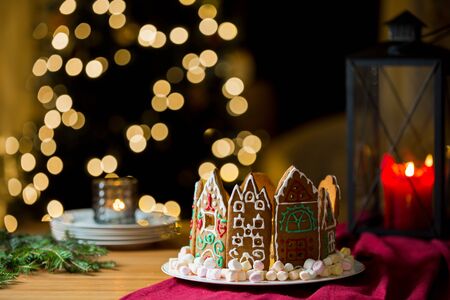 Little gingerbread houses with glaze standing on table with red tablecloth and decorations, candles and lanterns. Lights of Christmas tree on background.  Powdered sugar falling like snow flakesの写真素材