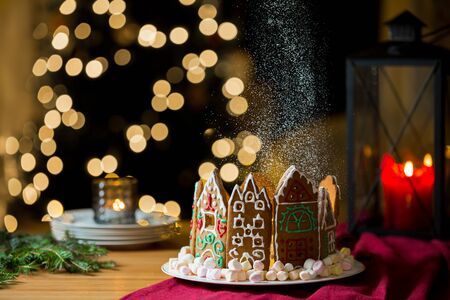Little gingerbread houses with glaze standing on table with red tablecloth and decorations, candles and lanterns. Lights of Christmas tree on background.  Powdered sugar falling like snow flakesの写真素材