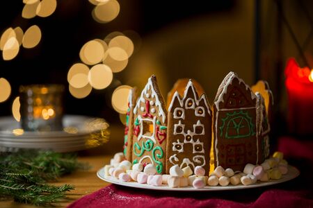 Little gingerbread houses with glaze standing on table with red tablecloth and decorations, candles and lanterns. Lights of Christmas tree on background.  Powdered sugar falling like snow flakesの写真素材