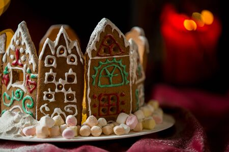 Little gingerbread houses with glaze standing on table with red tablecloth and decorations, candles and lanterns. Lights of Christmas tree on background.  Powdered sugar falling like snow flakesの写真素材