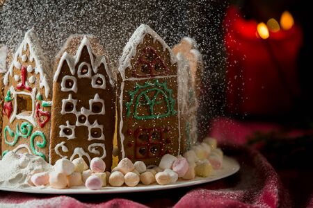 Little gingerbread houses with glaze standing on table with red tablecloth and decorations, candles and lanterns. Lights of Christmas tree on background.  Powdered sugar falling like snow flakesの写真素材