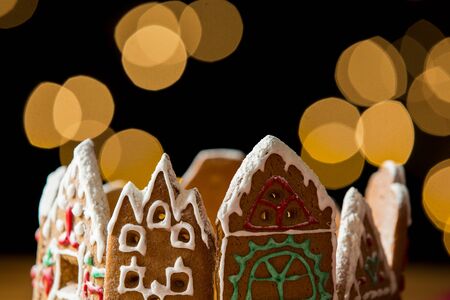 Little gingerbread houses with glaze standing on table with red tablecloth and decorations, candles and lanterns. Lights of Christmas tree on background.  Powdered sugar falling like snow flakesの写真素材