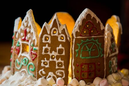 Little gingerbread houses with glaze standing on table with red tablecloth and decorations, candles and lanterns. Lights of Christmas tree on background.  Powdered sugar falling like snow flakesの写真素材