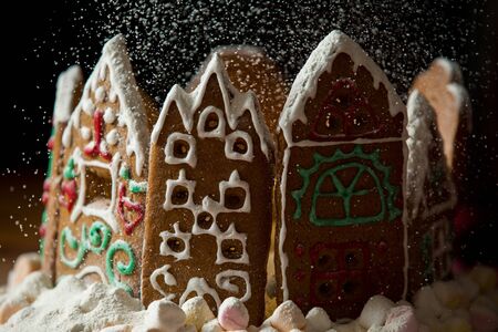 Little gingerbread houses with glaze standing on table with red tablecloth and decorations, candles and lanterns. Lights of Christmas tree on background.  Powdered sugar falling like snow flakesの写真素材