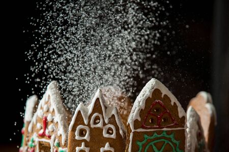 Little gingerbread houses with glaze standing on table with red tablecloth and decorations, candles and lanterns. Lights of Christmas tree on background.  Powdered sugar falling like snow flakesの写真素材