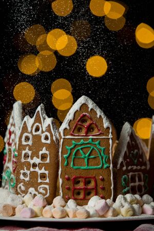 Little gingerbread houses with glaze standing on table with red tablecloth and decorations, candles and lanterns. Lights of Christmas tree on background.  Powdered sugar falling like snow flakesの写真素材