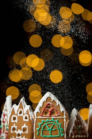 Little gingerbread houses with glaze standing on table with red tablecloth and decorations, candles and lanterns. Lights of Christmas tree on background.  Powdered sugar falling like snow flakesの写真素材