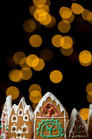 Little gingerbread houses with glaze standing on table with red tablecloth and decorations, candles and lanterns. Lights of Christmas tree on background.  Powdered sugar falling like snow flakesの写真素材