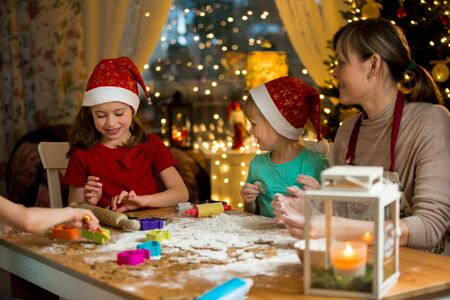 Mother and little kids in red hats cooking gingerbread cookies and playing. Beautiful living room with lights and Christmas tree, table with lantern. Happy family celebrating holiday together.の写真素材