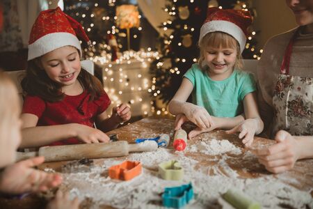 Mother and little kids in red hats cooking gingerbread cookies and playing. Beautiful living room with lights and Christmas tree, table with lantern. Happy family celebrating holiday together.の写真素材