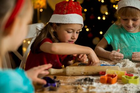 Mother and little kids in red hats cooking gingerbread cookies and playing. Beautiful living room with lights and Christmas tree, table with lantern. Happy family celebrating holiday together.の写真素材