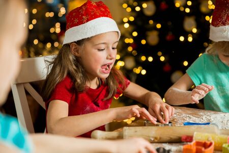 Mother and little kids in red hats cooking gingerbread cookies and playing. Beautiful living room with lights and Christmas tree, table with lantern. Happy family celebrating holiday together.の写真素材