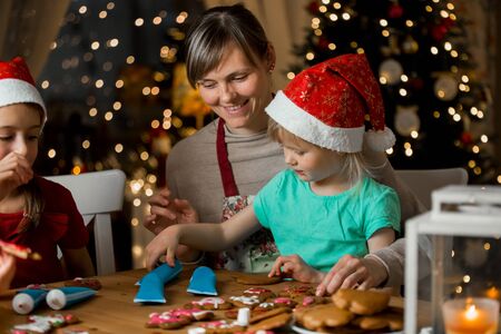 Mother and little kids in red hats cooking gingerbread cookies and decorating with glaze. Beautiful living room with lights and Christmas tree. Happy family celebrating holiday together.の写真素材