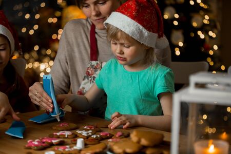 Mother and little kids in red hats cooking gingerbread cookies and decorating with glaze. Beautiful living room with lights and Christmas tree. Happy family celebrating holiday together.の写真素材