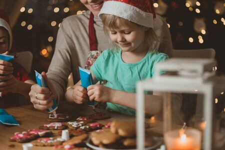 Mother and little kids in red hats cooking gingerbread cookies and decorating with glaze. Beautiful living room with lights and Christmas tree. Happy family celebrating holiday together.の写真素材