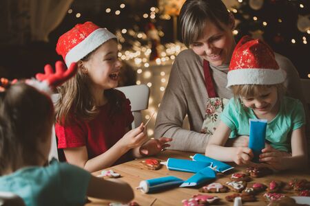 Mother and little kids in red hats cooking gingerbread cookies and decorating with glaze. Beautiful living room with lights and Christmas tree. Happy family celebrating holiday together.の写真素材