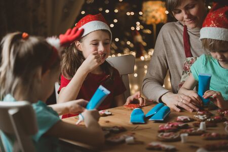 Mother and little kids in red hats cooking gingerbread cookies and decorating with glaze. Beautiful living room with lights and Christmas tree. Happy family celebrating holiday together.の写真素材