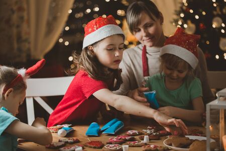 Mother and little kids in red hats cooking gingerbread cookies and decorating with glaze. Beautiful living room with lights and Christmas tree. Happy family celebrating holiday together.の写真素材