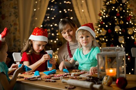 Mother and little kids in red hats cooking gingerbread cookies and decorating with glaze. Beautiful living room with lights and Christmas tree. Happy family celebrating holiday together.の写真素材