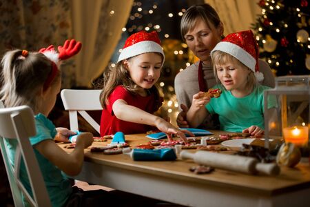 Mother and little kids in red hats cooking gingerbread cookies and decorating with glaze. Beautiful living room with lights and Christmas tree. Happy family celebrating holiday together.の写真素材