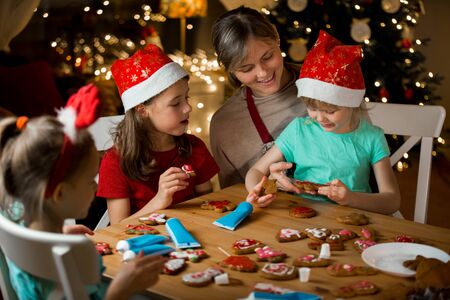 Mother and little kids in red hats cooking gingerbread cookies and decorating with glaze. Beautiful living room with lights and Christmas tree. Happy family celebrating holiday together.の写真素材