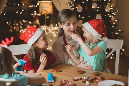 Mother and little kids in red hats cooking gingerbread cookies and decorating with glaze. Beautiful living room with lights and Christmas tree. Happy family celebrating holiday together.の写真素材