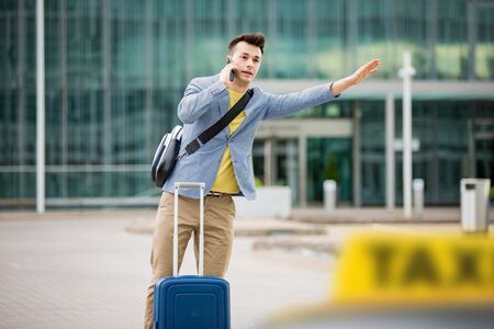 Stylish man standing at airport with suitcase and smartphone, hailing taxi.の写真素材