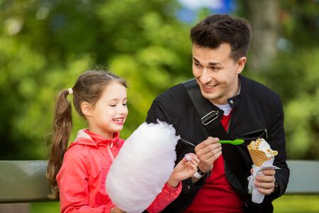 Cute girl and father eating cotton candy and ice cream, sitting on bench in park. Happy family having quality time togetherの写真素材