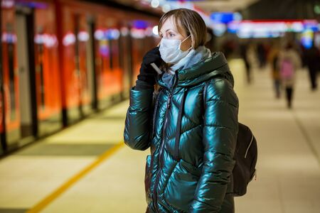 Woman in winter coat with protective mask on face standing on metro station, waiting for train, looking worried. Preventive measures in public places of epidemic regions. Finland, Espooの写真素材