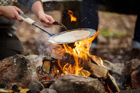 Man and woman making pancakes on campfire in forest on shore of lake, making a fire, grilling. Happy couple exploring Finland. Scandinavian landscape.の写真素材