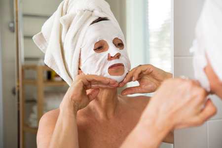 Mature beautiful woman taking off facial sheet mask. Looking in the mirror in bathroom, Wrapped in a towel. Skincare routine and anti-aging treatment at home for older women.の写真素材