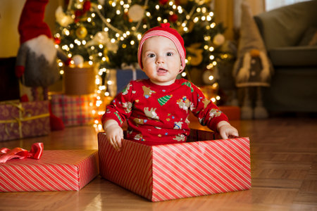 Happy baby boy in a red cap sitting in a gift box on the background of a Christmas tree on Christmas Eve. Happy Holidays, New Year. Cozy warm winter evening at home. Xmas timeの写真素材