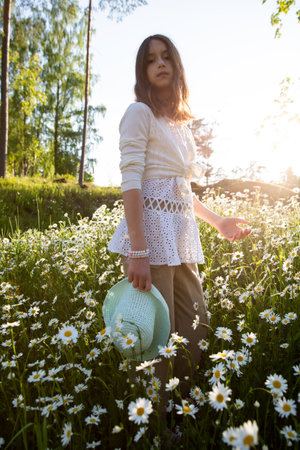 Girl standing on a meadow covered with wild flowers. Holding hat on hand. Background of field of daisy flowers in bloom. Sunny summer day, rays of light. Summer vacation conceptの写真素材