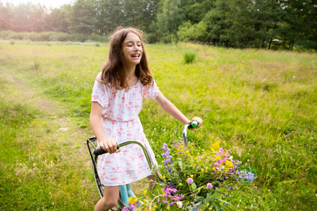 Girl on a bike with a basket of wild flowers on a meadow. Sunny summer day, rays of light, rural background. Summer vacation concept.の写真素材